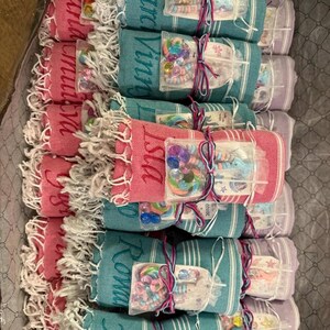 May include: A collection of rolled-up beach towels in various colors, including pink, teal, and lavender, with white fringe. Each towel is tied with a ribbon and includes a small package of colorful hair accessories. Some towels have names embroidered on them.