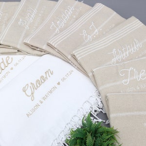 May include: A collection of personalized beige and white towels, embroidered with names and wedding details. The towels feature names like "Bride," "Groom," and other names, with dates and names embroidered below. The towels have a striped design.