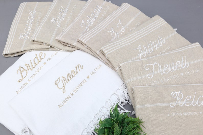 May include: A collection of personalized beige and white towels, embroidered with names and wedding details. The towels feature names like "Bride," "Groom," and other names, with dates and names embroidered below. The towels have a striped design.