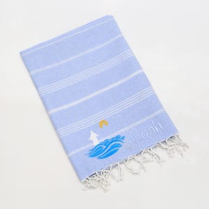 May include: A light blue beach towel with white stripes and a fringed edge. The towel features an embroidered design of a lighthouse, waves, and the name "Noah".