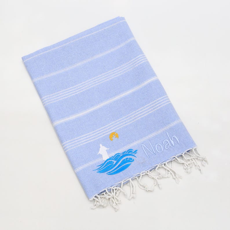 May include: A light blue beach towel with white stripes and a fringed edge. The towel features an embroidered design of a lighthouse, waves, and the name "Noah".