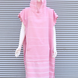 May include: A pink hooded poncho towel featuring thin horizontal white stripes, short sleeves, and a large front kangaroo pocket. The garment has a straight hemline finished with decorative white fringe tassels along the bottom edge.