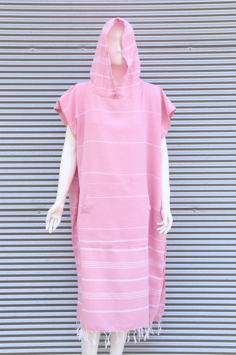 May include: A pink hooded poncho towel featuring thin horizontal white stripes, short sleeves, and a large front kangaroo pocket. The garment has a straight hemline finished with decorative white fringe tassels along the bottom edge.
