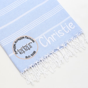 May include: A light blue and white striped beach towel with white fringe. The towel has the name "Christie" embroidered in white. A circular emblem with text and a symbol is also embroidered on the towel.