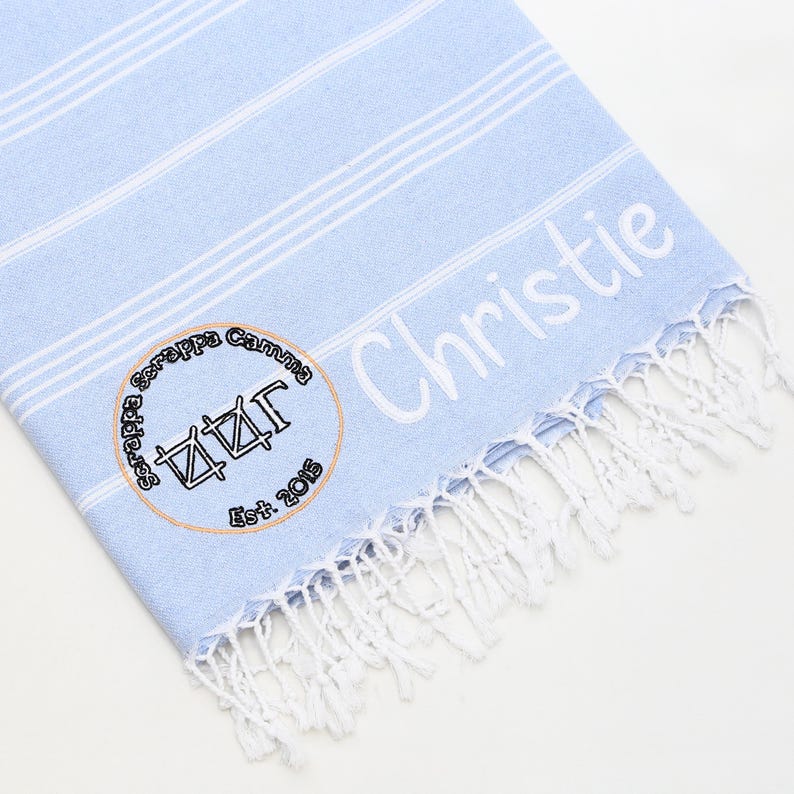 May include: A light blue and white striped beach towel with white fringe. The towel has the name "Christie" embroidered in white. A circular emblem with text and a symbol is also embroidered on the towel.