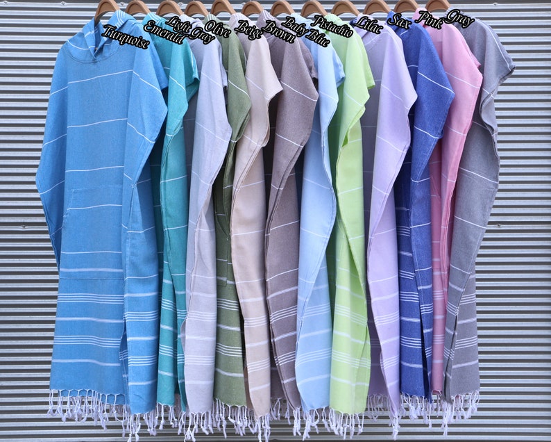 May include: A collection of hooded poncho towels with fringe trim, featuring horizontal white stripes. Colors include turquoise, emerald, light gray, olive, beige, brown, baby blue, pistachio, lilac, sax, pink, and gray, each labeled with its color name.
