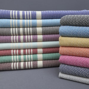 Könnte beinhalten: Ein Stapel von 15 türkischen Handtüchern in verschiedenen Farben, darunter Blau, Schwarz, Lila, Rot, Grün, Orange, Pink und Weiß. Die Handtücher haben ein Fischgrätenmuster und Fransen an den Enden.