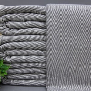 May include: A stack of grey and white herringbone patterned throw blankets with white fringe. The blankets are folded and stacked on a grey surface.