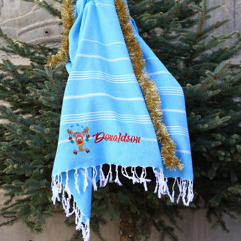 May include: A light blue beach towel with white stripes and a fringed edge. The towel features an embroidered reindeer design and the name "Donaldson" in red. Gold tinsel decorates the scene.