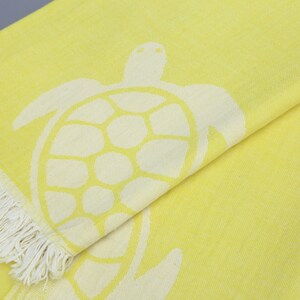 Op de afbeelding: Een gele strandhanddoek met een wit zeeschildpad ontwerp. De handdoek heeft een franje rand.