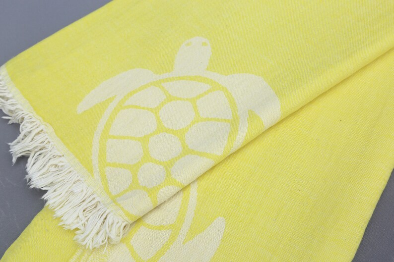 Op de afbeelding: Een gele strandhanddoek met een wit zeeschildpad ontwerp. De handdoek heeft een franje rand.