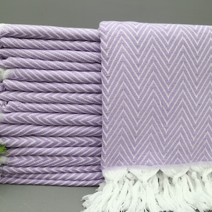 May include: A stack of lavender and white chevron-patterned towels with white fringe. The towels are neatly folded, showcasing the intricate zigzag design. The soft color palette and texture suggest a spa or bathroom theme.