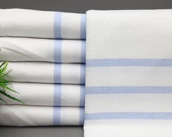 Può includere: Un mucchio di asciugamani bianchi con strisce blu chiaro. Gli asciugamani sono piegati e disposti in una pila ordinata.