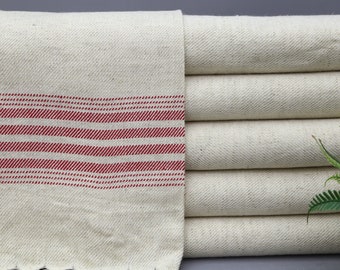 Puede incluir: Una pila de cinco toallas de cocina de algodón de color blanco roto con rayas rojas. Las toallas están dobladas y apiladas sobre una superficie gris.
