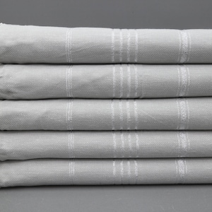 May include: A stack of six grey Turkish towels with white fringe. The towels are folded and stacked on a grey surface.