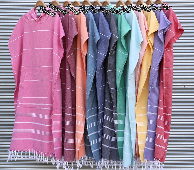May include: A collection of hooded poncho towels hanging on wooden hangers, each featuring a front pocket, white horizontal stripes, and a fringed hem. The colors include fuchsia, burgundy, orange, teal, navy, black, green, mint, salmon, yellow, purple, and red.