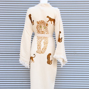 May include: A white cotton robe with a fringe trim and a leopard print design. The robe has a belt and features several leopards in different poses.