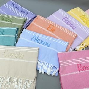 May include: A collection of colorful, personalized Turkish towels. Each towel features embroidered names in various colors, including blue, pink, green, and yellow. The towels have white fringe and striped details, perfect for beach or bath use.