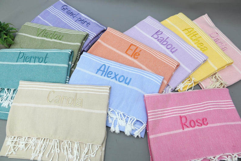 May include: A collection of colorful, personalized Turkish towels. Each towel features embroidered names in various colors, including blue, pink, green, and yellow. The towels have white fringe and striped details, perfect for beach or bath use.