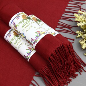 May include: Two burgundy pashmina scarves with fringed edges, rolled up and tied with white labels that say "Stay Warm" and include the date "12 March 2024" and the words "Name & Name".