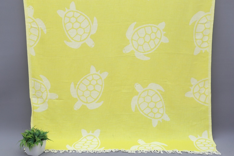 Op de afbeelding: Een gele strandhanddoek met een wit schildpadpatroon. De handdoek heeft een gefranst randje.