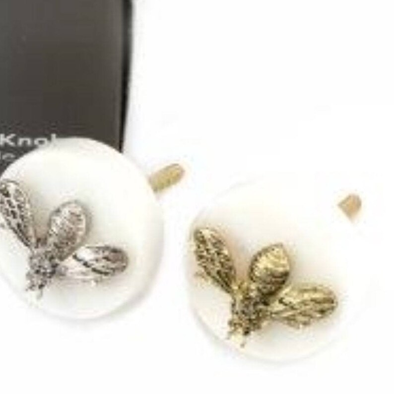 May include: Two white ceramic drawer knobs with a gold bee design. The bee is detailed with textured wings and a body.
