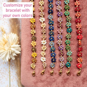Op de afbeelding: Zeven kralenarmbanden met bloemvormige kralen in verschillende kleuren, waaronder geel, bruin, crème, blauw, paars, groen en rood. De armbanden liggen op een roze stoffen oppervlak. Personaliseer je armband met je eigen kleuren.