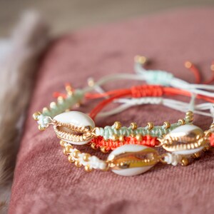 Peut inclure: Trois bracelets avec des coquillages blancs et des accents dorés. Les bracelets sont faits de ficelle rouge, blanche et verte.