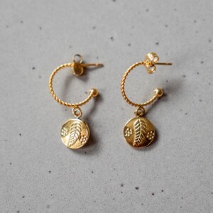 Op de afbeelding: Een paar gouden creolen met een bladhanger. De oorbellen zijn gemaakt van een dunne, gedraaide draad en hebben een eenvoudig, elegant ontwerp.