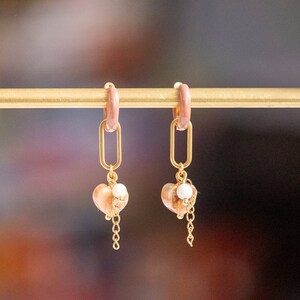 Op de afbeelding: Een paar gouden creolen met een roze harsring en een hangende hartbedel. De hartbedel is gemaakt van een lichtbruine schelp met een witte parel accent en een gouden ketting.