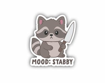 May include: A die-cut sticker featuring a cartoon raccoon with gray and brown fur, holding a kitchen knife in one paw. The raccoon has a serious facial expression. Below the illustration, the text reads MOOD: STABBY in brown capital letters.