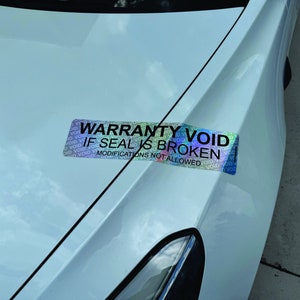 May include: A holographic sticker with the text "WARRANTY VOID IF SEAL IS BROKEN MODIFICATIONS NOT ALLOWED" is affixed to the white hood of a car. The sticker has a rainbow sheen and the word "VOID" is repeated.