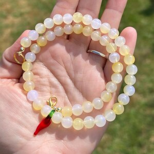 May include: A beaded necklace with yellow and white round beads. A red chili pepper charm with green accents hangs from the necklace. The necklace is held in a hand against a blurred green background.