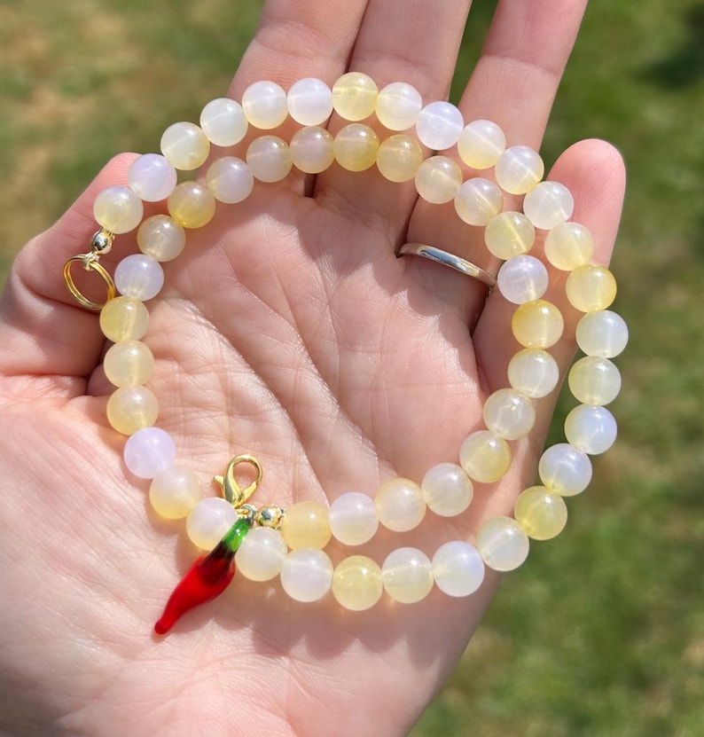 May include: A beaded necklace with yellow and white round beads. A red chili pepper charm with green accents hangs from the necklace. The necklace is held in a hand against a blurred green background.