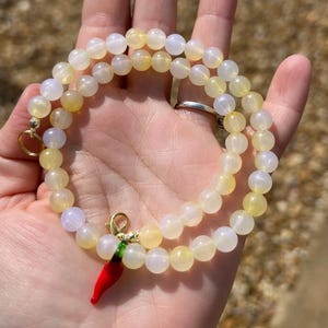 May include: A beaded necklace with yellow and white round beads. The necklace has a gold clasp and a red chili pepper charm with green accents. The necklace is held in a hand.