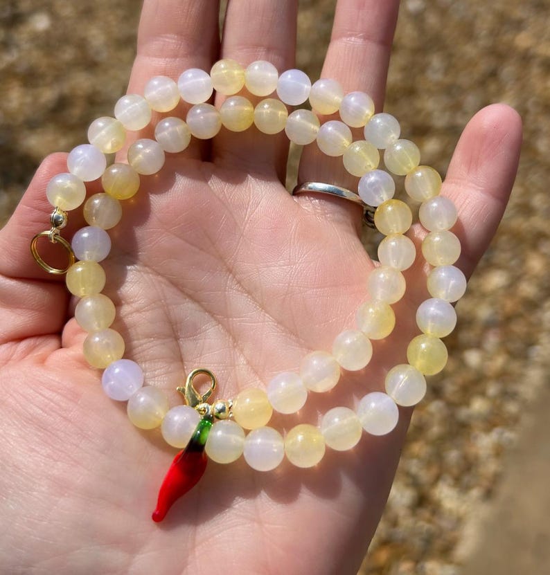 May include: A beaded necklace with yellow and white round beads. The necklace has a gold clasp and a red chili pepper charm with green accents. The necklace is held in a hand.