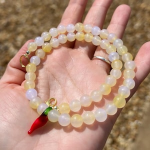 May include: A beaded necklace with yellow and white beads. A red chili pepper charm with green accents hangs from the necklace. The necklace is displayed on a hand.