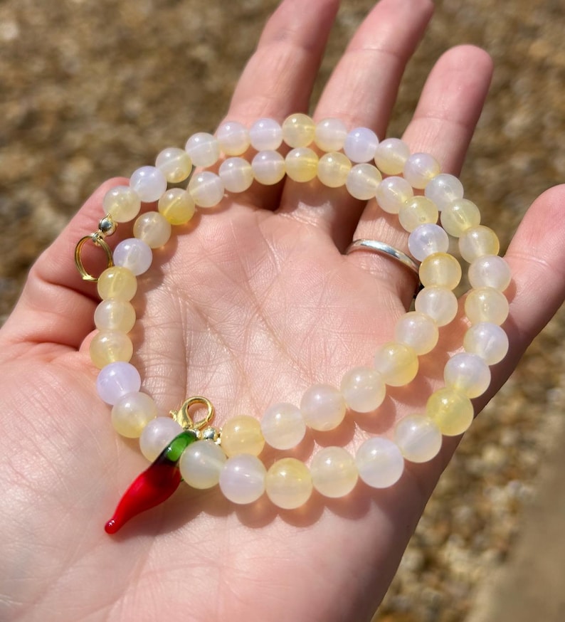 May include: A beaded necklace with yellow and white beads. A red chili pepper charm with green accents hangs from the necklace. The necklace is displayed on a hand.