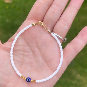 May include: White beaded bracelet with a gold clasp and a blue evil eye charm.