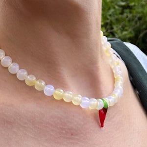May include: A beaded necklace with a pendant. The necklace features round, off-white and yellow beads. A red chili pepper pendant hangs from the necklace. The necklace is worn around the neck.