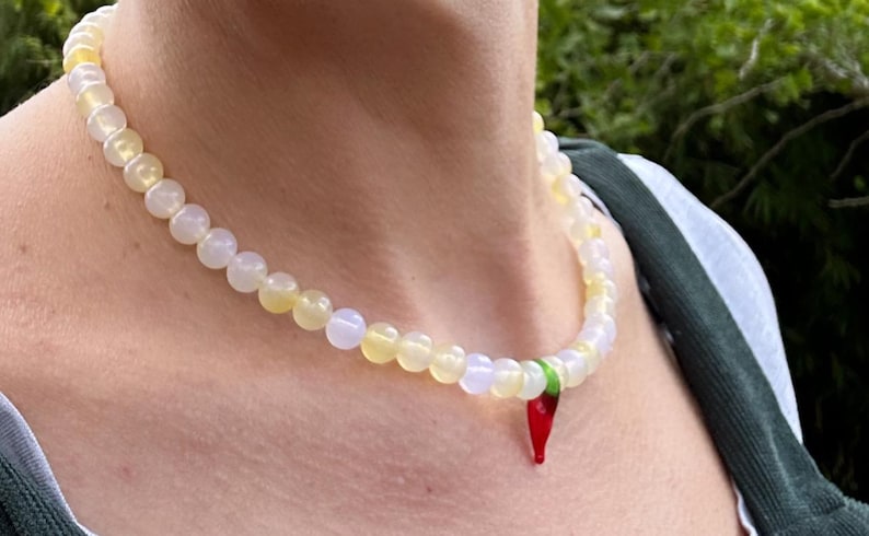 May include: A beaded necklace with a pendant. The necklace features round, off-white and yellow beads. A red chili pepper pendant hangs from the necklace. The necklace is worn around the neck.