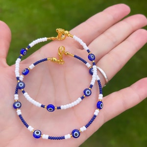 May include: Two beaded bracelets with blue evil eye charms and white and blue beads. The bracelets have gold clasps.