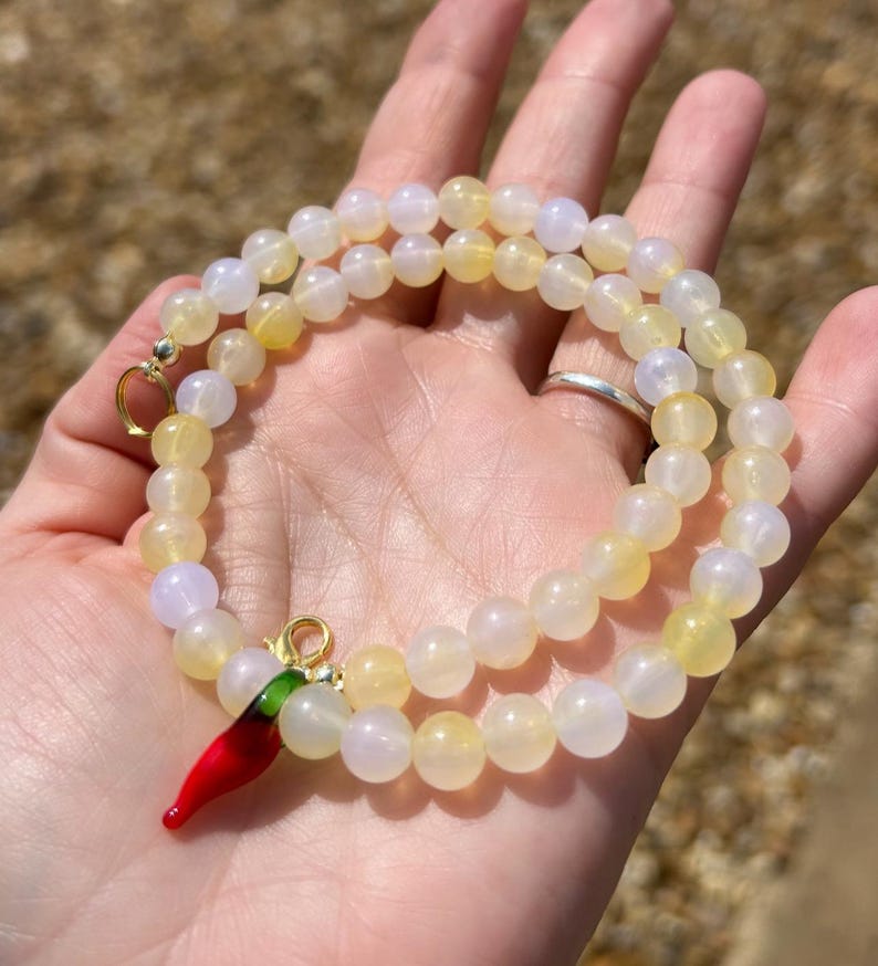 May include: A beaded necklace with yellow and white beads. A red chili pepper charm with green accents hangs from the necklace. The necklace is displayed on a hand.