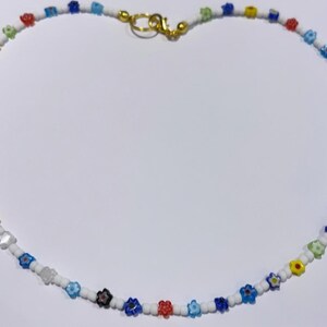 Puede incluir: Un collar de cuentas blancas con cuentas de colores en forma de flor. El collar tiene un cierre dorado.