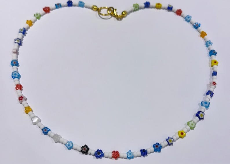 Puede incluir: Un collar de cuentas blancas con cuentas de colores en forma de flor. El collar tiene un cierre dorado.