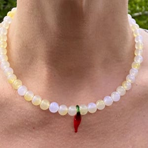 May include: A beaded necklace with a pendant. The necklace features small, round, off-white and yellow beads. A red chili pepper pendant with green accents hangs from the center of the necklace. The necklace is worn around the neck.