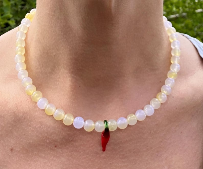 May include: A beaded necklace with a pendant. The necklace features small, round, off-white and yellow beads. A red chili pepper pendant with green accents hangs from the center of the necklace. The necklace is worn around the neck.