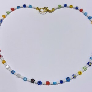 Puede incluir: Un collar de cuentas de colores con peque&ntilde;as cuentas con forma de flor en tonos de azul, rojo, amarillo y blanco. El collar est&aacute; hecho con cuentas blancas y tiene un cierre dorado.