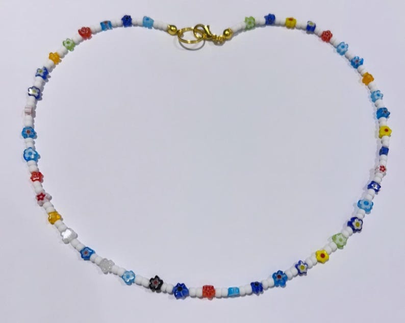 Puede incluir: Un collar de cuentas de colores con peque&ntilde;as cuentas con forma de flor en tonos de azul, rojo, amarillo y blanco. El collar est&aacute; hecho con cuentas blancas y tiene un cierre dorado.