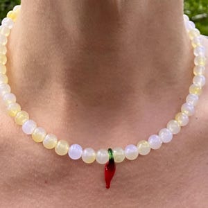 May include: A beaded necklace with a pendant. The necklace features a strand of round, light yellow and white beads. A small, red chili pepper pendant with green accents hangs from the center of the necklace.
